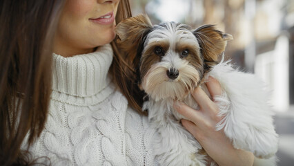 A young hispanic woman happily holds her beautiful biewer yorkshire terrier on a bustling city...