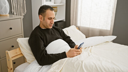 Fototapeta premium Handsome hispanic man relaxing in bedroom using smartphone, conveying modern indoor lifestyle