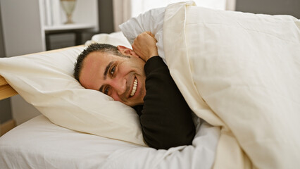 Fototapeta na wymiar A smiling young man nestled in bed sheets expressing comfort and relaxation in a cozy bedroom setting.