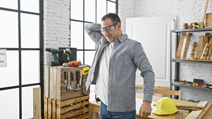 A hispanic man expressing back pain in a well-organized carpentry workshop with various tools and a...