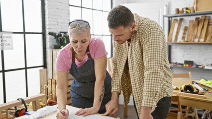 A man and woman team up as carpenters analyzing plans in a woodworking workshop, embodying craftsmanship and collaboration.