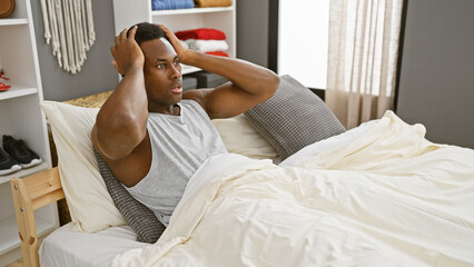 A concerned young african american man sitting in bed within a modern bedroom interior, conveying a...