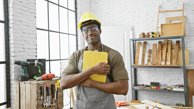 A confident young african american man wearing a hard hat and safety glasses stands in a well-equipped carpentry workshop holding a clipboard.