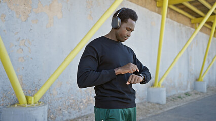 A young african american man checks his smartwatch while listening to music with headphones in an urban outdoor setting.