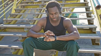 A fit african american man rests on urban stairs wearing headphones and checking his fitness tracker in a sunlit city environment.