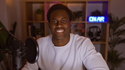 A smiling young adult african american man with a microphone in a cozy room illuminated by neon 'on...