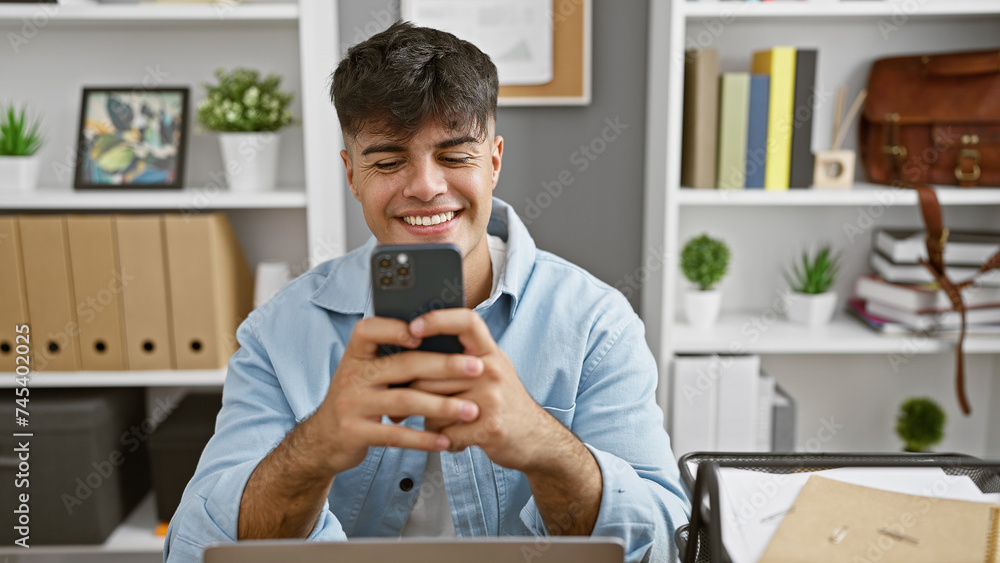 Poster Handsome young hispanic man happily working on his business at the office, enjoying typing a message on his smartphone, confidently smiling in a professional setting indoors