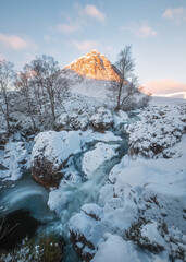 Winter waterfall at the foot of a snow-capped mountain with a morning sunlit peak. Etive Mor Waterfall. Scottish Highlands, Scotland