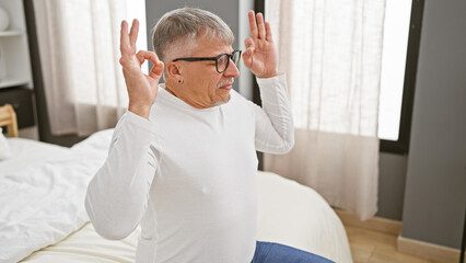 Fototapeta premium A middle-aged man with grey hair, wearing glasses, makes gestures sitting on a bed, indoors, portraying a relaxed or playful mood.