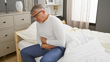 Fototapeta premium A mature man in distress sitting on bed clutching his stomach in a well-furnished bedroom.