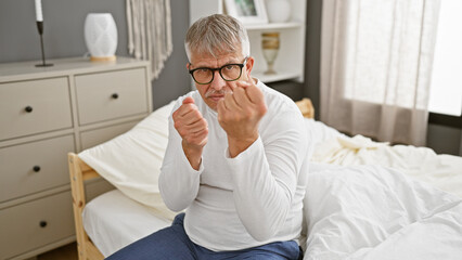 Mature grey-haired man in glasses making fists while sitting on a bed in a well-lit bedroom