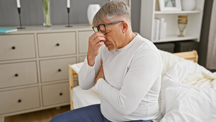 Mature grey-haired man in glasses feeling unwell, sitting on a bed in a modern bedroom.