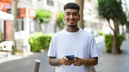 Smiling young latin man happily texting on his smartphone outdoors, a handsome tattooed adult immersed in a cheerful digital conversation on the urban street. - obrazy, fototapety, plakaty