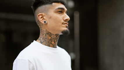 Cool, tattooed young latin man giving a serious look, outdoors. a relaxed pose on a sunny city street, concentrating on his urban lifestyle. - obrazy, fototapety, plakaty