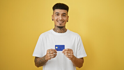 Cheerful young hispanic man exuding confidence, smiling with credit card against a yellow isolated background