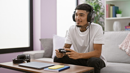 Cheerful young latin man sitting comfortably on a sofa at home, engrossed in playing a gripping video game with wireless control, exuding confidence with a charming smile.