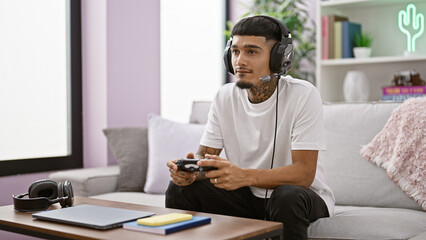 Handsome young latin man, tattooed and serious, sitting comfortably on his home sofa, deeply immersed in the world of online gaming using a controller, headphones at the ready.
