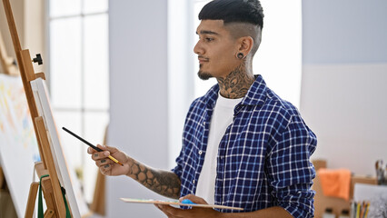In an indoor art studio, a young latin artist with tattoos concentrates deeply, thinking and...