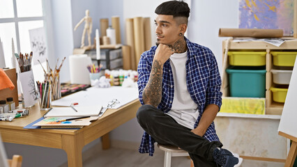 Tattooed young latin man, an engrossed artist, sitting at his easel with serious face in an art...