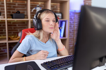 Young caucasian woman playing video games wearing headphones hand on mouth telling secret rumor, whispering malicious talk conversation - obrazy, fototapety, plakaty