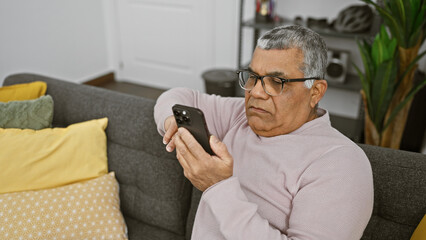 Mature grey-haired man in glasses using smartphone at home, expresses contemplation.