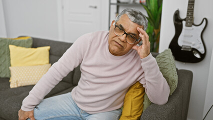 Portrait of a pensive senior man with glasses, sitting on a couch indoors with a guitar in the blurry background.
