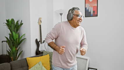 Grey-haired man dances joyfully at home wearing headphones, showcasing a cheerful senior lifestyle.