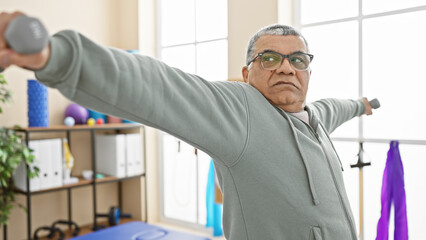 Senior man exercising with dumbbells in a rehab clinic indoors, focused on maintaining health and...