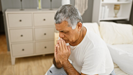 A pensive mature man with grey hair sitting in a bedroom, contemplating or praying with clasped hands.