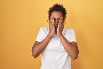 Young hispanic woman with curly hair standing over yellow background rubbing eyes for fatigue and...