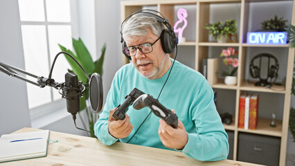 A grey-haired man examines game controllers while recording in an indoor studio with an 'on air'...