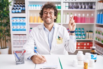 Hispanic young man working at pharmacy drugstore gesturing finger crossed smiling with hope and eyes closed. luck and superstitious concept.
