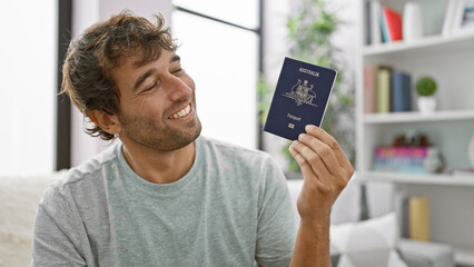 Cheerful, confident young man, casually sitting on his living room sofa, holding proudly the passport of australia and smiling joyfully.