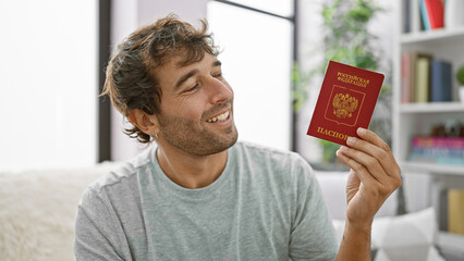 Cheery young man tours his living room, blissfully holding high a russian passport, relaxed sitting...