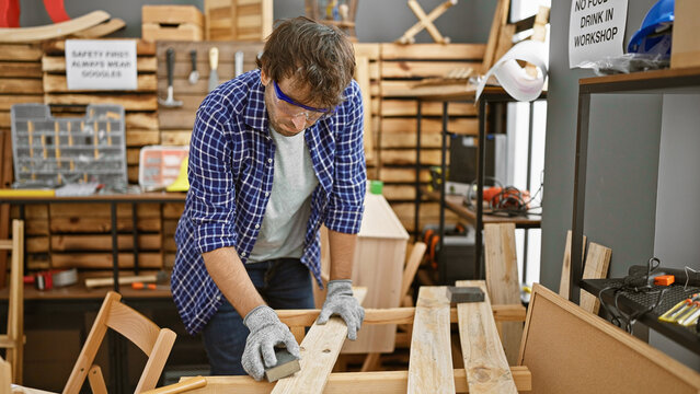 Handsome, young, bearded man with glasses, engrossed in his profession, sanding a wooden plank at a bustling carpentry workshop