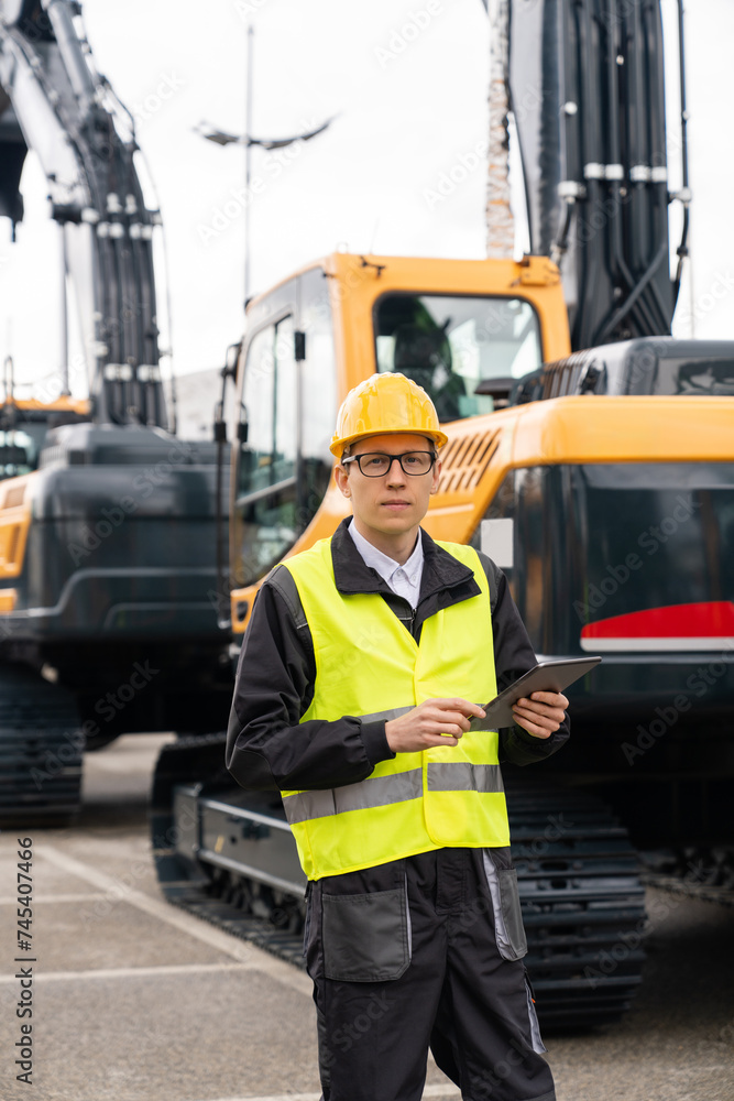 Poster Engineer in a helmet with a digital tablet stands next to construction excavators..