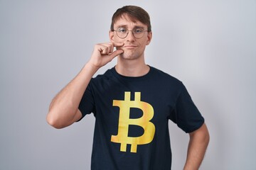 Caucasian blond man wearing bitcoin t shirt mouth and lips shut as zip with fingers. secret and...