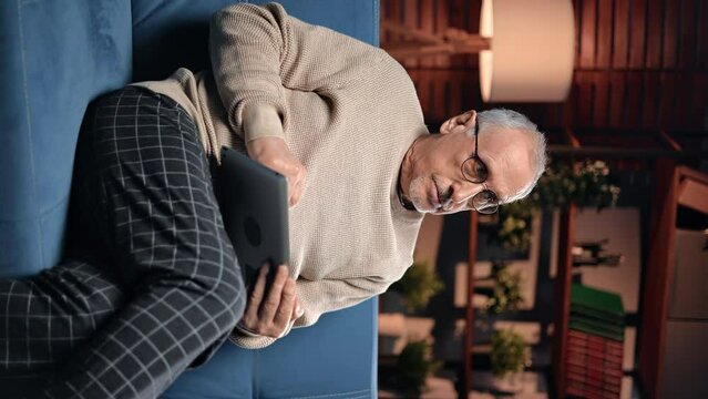 VERTICAL VIDEO POV Portrait of elderly man with modern tablet at home in evening, surfing internet