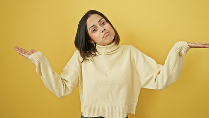 A perplexed young hispanic woman shrugs against a yellow background, showing uncertainty and...