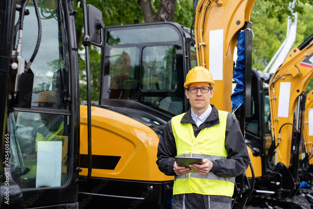 Poster engineer in a helmet with a digital tablet stands next to construction excavators..