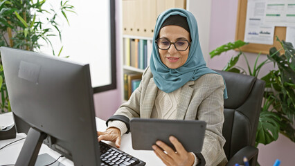 Professional woman wearing hijab multitasking with a tablet and computer in a modern office setting.