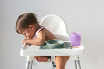 unhappy baby sitting in a high chair on a white background, eating food, all dirty and tear-stained