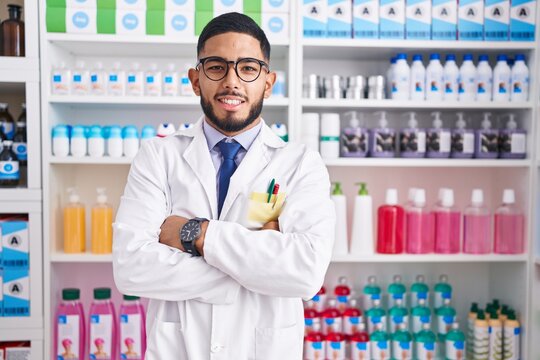 Young latin man pharmacist smiling confident standing with arms crossed gesture at pharmacy