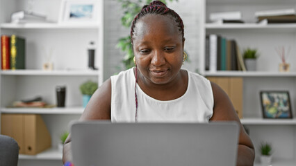 Hard-working african american woman, boss with braids, focused on her laptop at the office, epitomizing professional success.