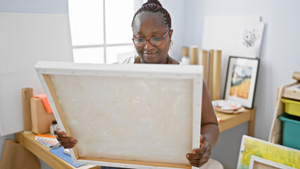 Confident african american woman artist smiling, enjoying her hobby, holding brush and painting on canvas while looking at her artwork in studio - obrazy, fototapety, plakaty
