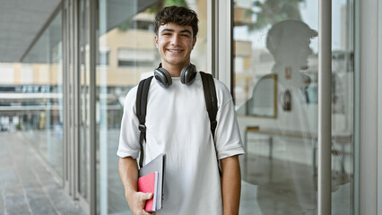 Confident young hispanic teenager, a joyful student wearing headphones, happily holding his university books, flashing a handsome, cheerful smile while casually standing on the lively city campus.