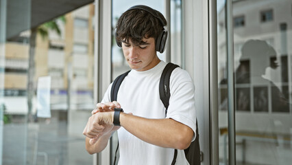 Cool young hispanic student, jamming to his favorite song on headphones, glances at his watch amid...