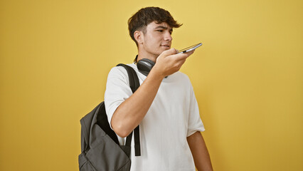 Tech-savvy young hispanic teenager, a portrait of a smart student sending a voice message online, standing isolated on a yellow background with his smartphone and headphones