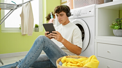 Cool young hispanic teenager engrossed in music, using touchpad and headphones while waiting for...