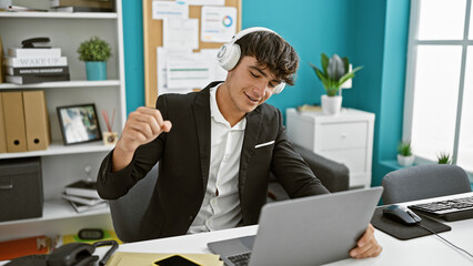 Handsome young hispanic teenager joyfully dancing around the office, listening to music via headphones while handling his business work on a laptop, spreading positive vibes. - obrazy, fototapety, plakaty